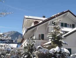Ein charmantes Haus mit einem schneebedeckten Dach und grünen Fensterläden. Im Hintergrund sind schneebedeckte Berge und ein strahlend blauer Himmel zu sehen.