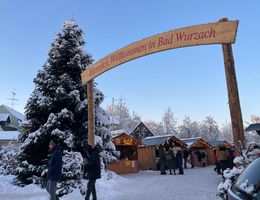 Im Vordergrund ein hölzerner Torbogen mit der Aufschrift "Herzlich Willlkommen in Bad Wurzach". Links daneben ein beschneiter Weihnachtsbaum und im Hintergrund der beschneite Klosterplatz mit hölzernen beleuchteten Hüttchen.