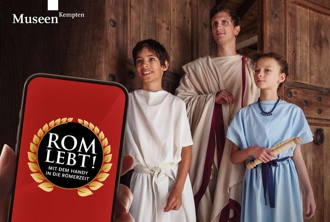 Drei als Römer der Antike gewandete Personen - ein Erwachsener und ein Junge und ein Mädchen- posieren in einer geöffneten Holztüre für ein Handy, das im Vordergrund sichtbar ist und den Namen der Ausstellung auf dem Display trägt
