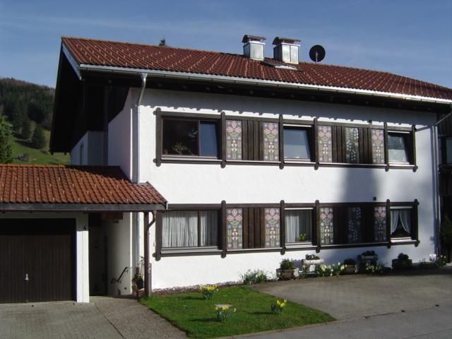 A two-story house with a red tiled roof and white walls. The windows are decorated and there is a small garden in the foreground.