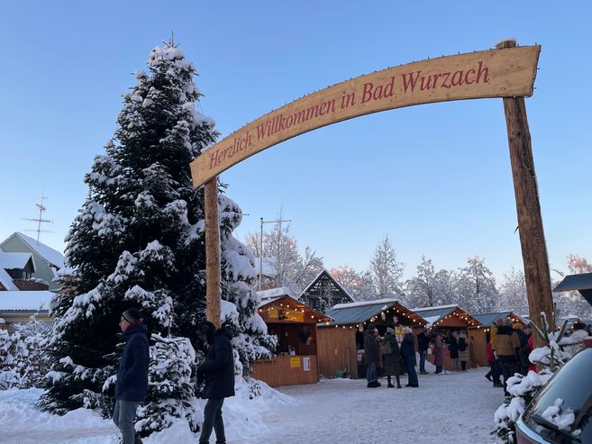 Im Vordergrund ein hölzerner Torbogen mit der Aufschrift "Herzlich Willlkommen in Bad Wurzach". Links daneben ein beschneiter Weihnachtsbaum und im Hintergrund der beschneite Klosterplatz mit hölzernen beleuchteten Hüttchen.