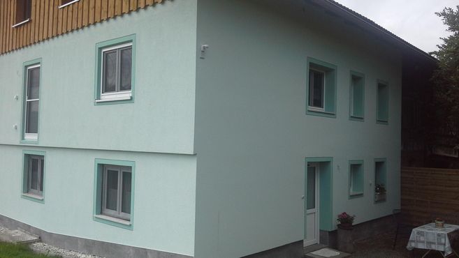 A modern two-story building with a light blue facade and white windows. In front of the house, there is a table with chairs.