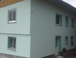 A modern two-story building with a light blue facade and white windows. In front of the house, there is a table with chairs.