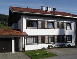 A two-story house with a red tiled roof and white walls. The windows are decorated and there is a small garden in the foreground.