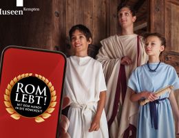 Drei als Römer der Antike gewandete Personen - ein Erwachsener und ein Junge und ein Mädchen- posieren in einer geöffneten Holztüre für ein Handy, das im Vordergrund sichtbar ist und den Namen der Ausstellung auf dem Display trägt