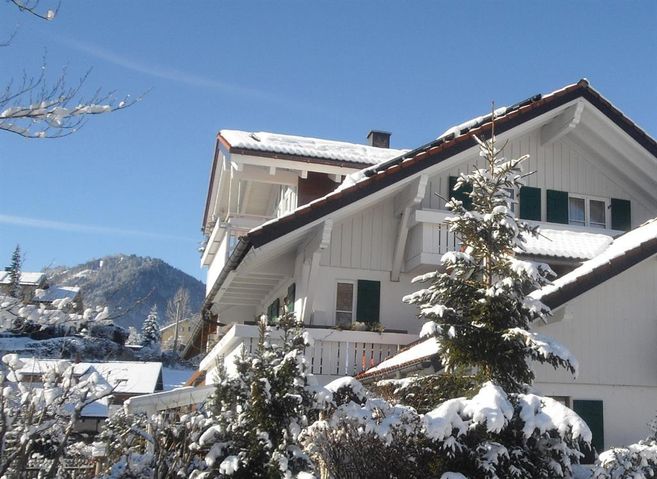 Ein charmantes Haus mit einem schneebedeckten Dach und grünen Fensterläden. Im Hintergrund sind schneebedeckte Berge und ein strahlend blauer Himmel zu sehen.